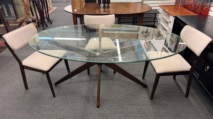 A mid-century modern dining set consisting of a round glass-top table on an angled dark-wood base paired with four curved-back off-white upholstered chairs in good condition.