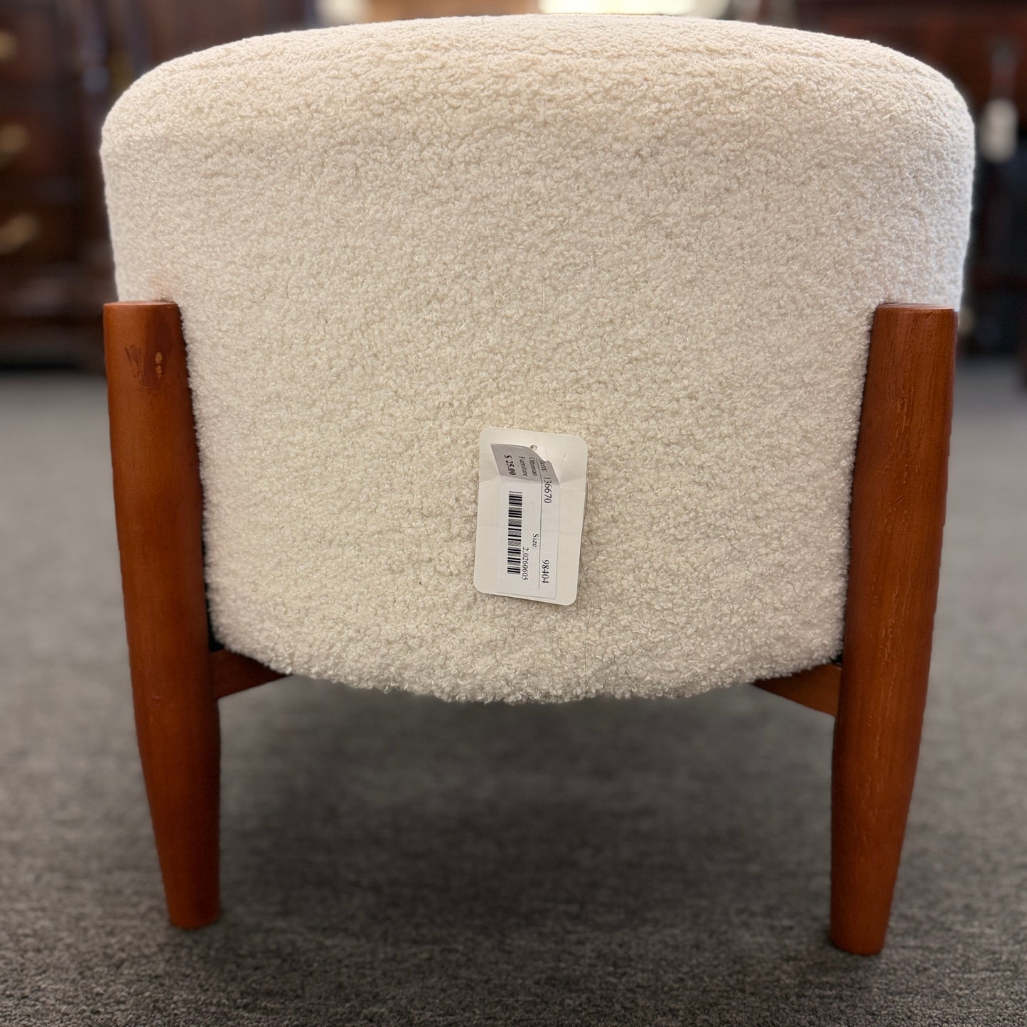 A mid-century modern round accent ottoman featuring a plush, fabric-upholstered cylindrical seat on a structured, four-legged exposed wooden base.