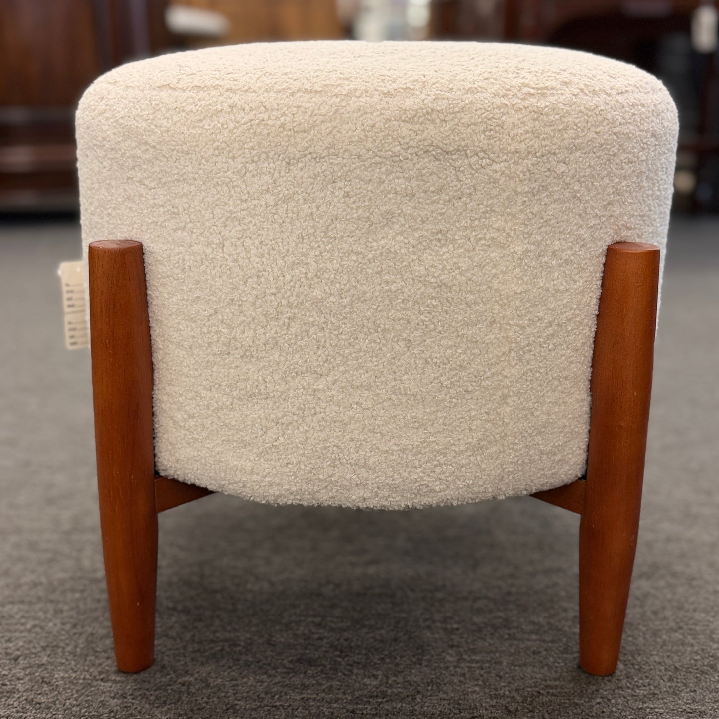 A mid-century modern round accent ottoman featuring a plush, fabric-upholstered cylindrical seat on a structured, four-legged exposed wooden base.