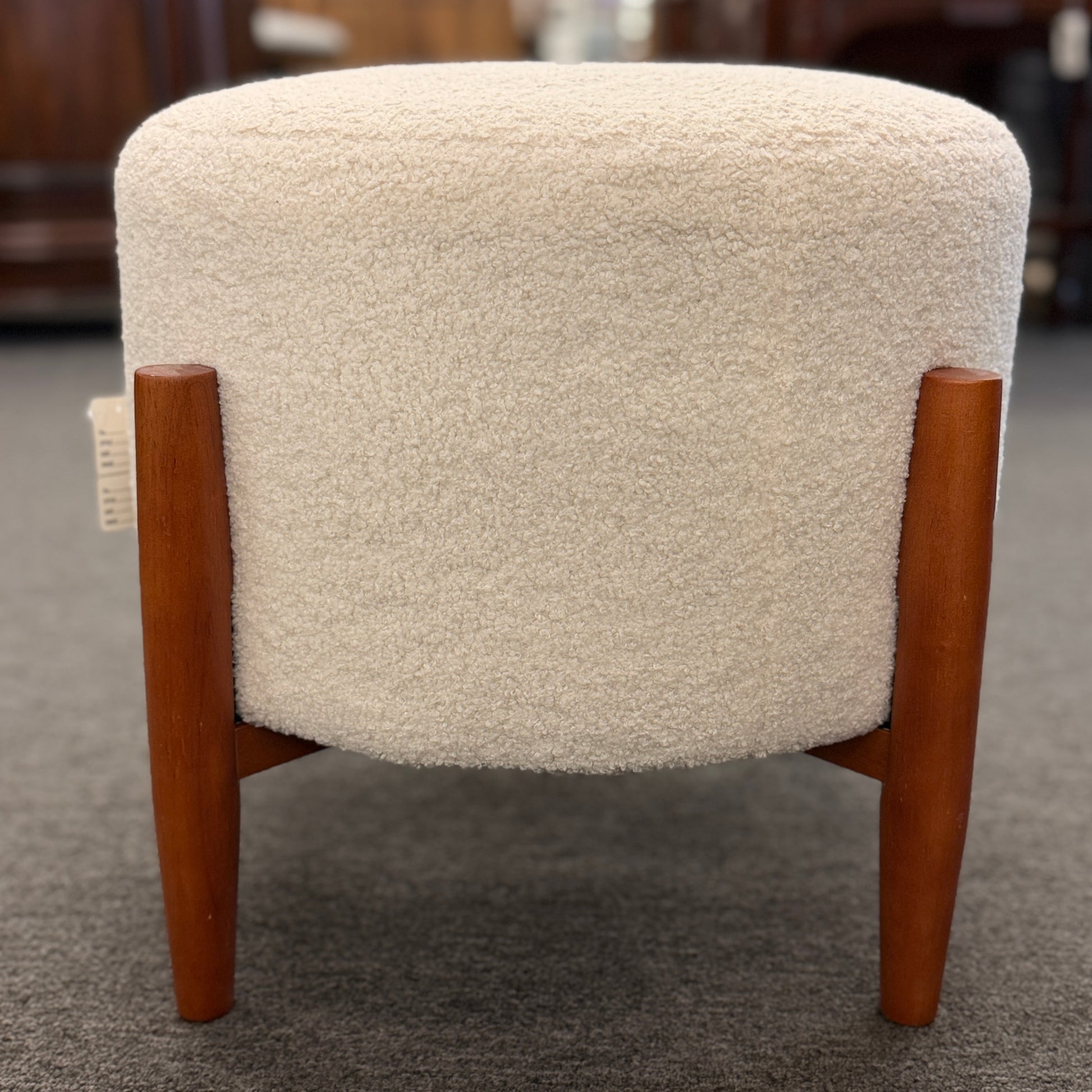 A mid-century modern round accent ottoman featuring a plush, fabric-upholstered cylindrical seat on a structured, four-legged exposed wooden base.