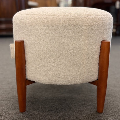 A mid-century modern round accent ottoman featuring a plush, fabric-upholstered cylindrical seat on a structured, four-legged exposed wooden base.