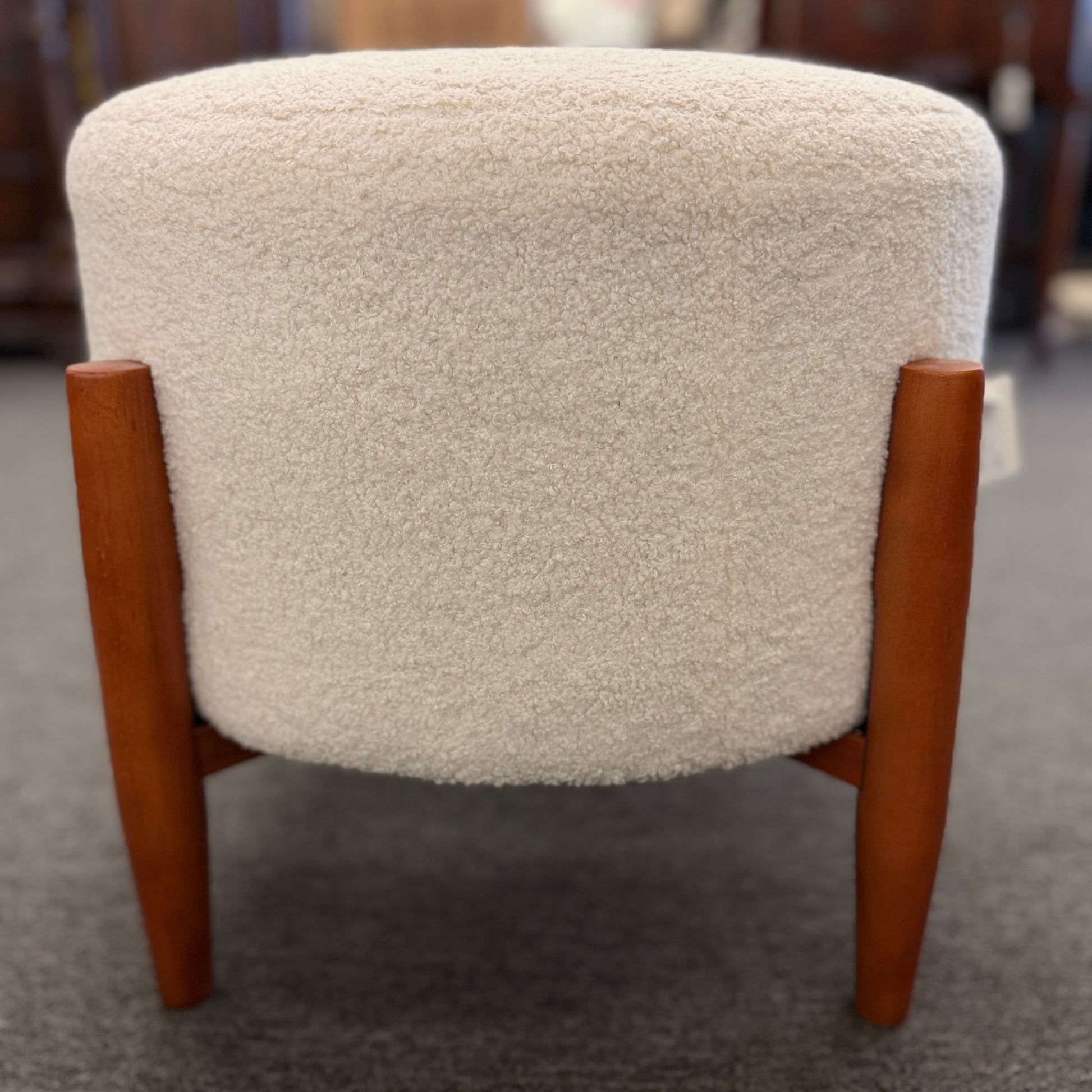 A mid-century modern round accent ottoman featuring a plush, fabric-upholstered cylindrical seat on a structured, four-legged exposed wooden base.