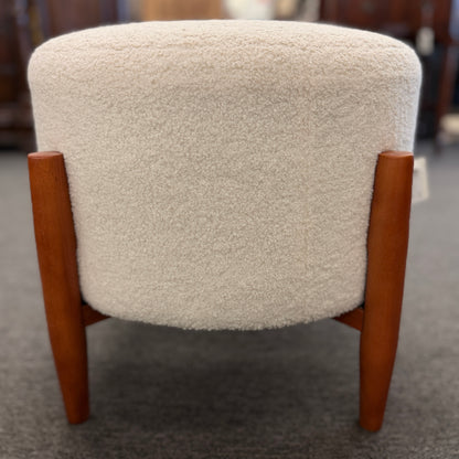 A mid-century modern round accent ottoman featuring a plush, fabric-upholstered cylindrical seat on a structured, four-legged exposed wooden base.