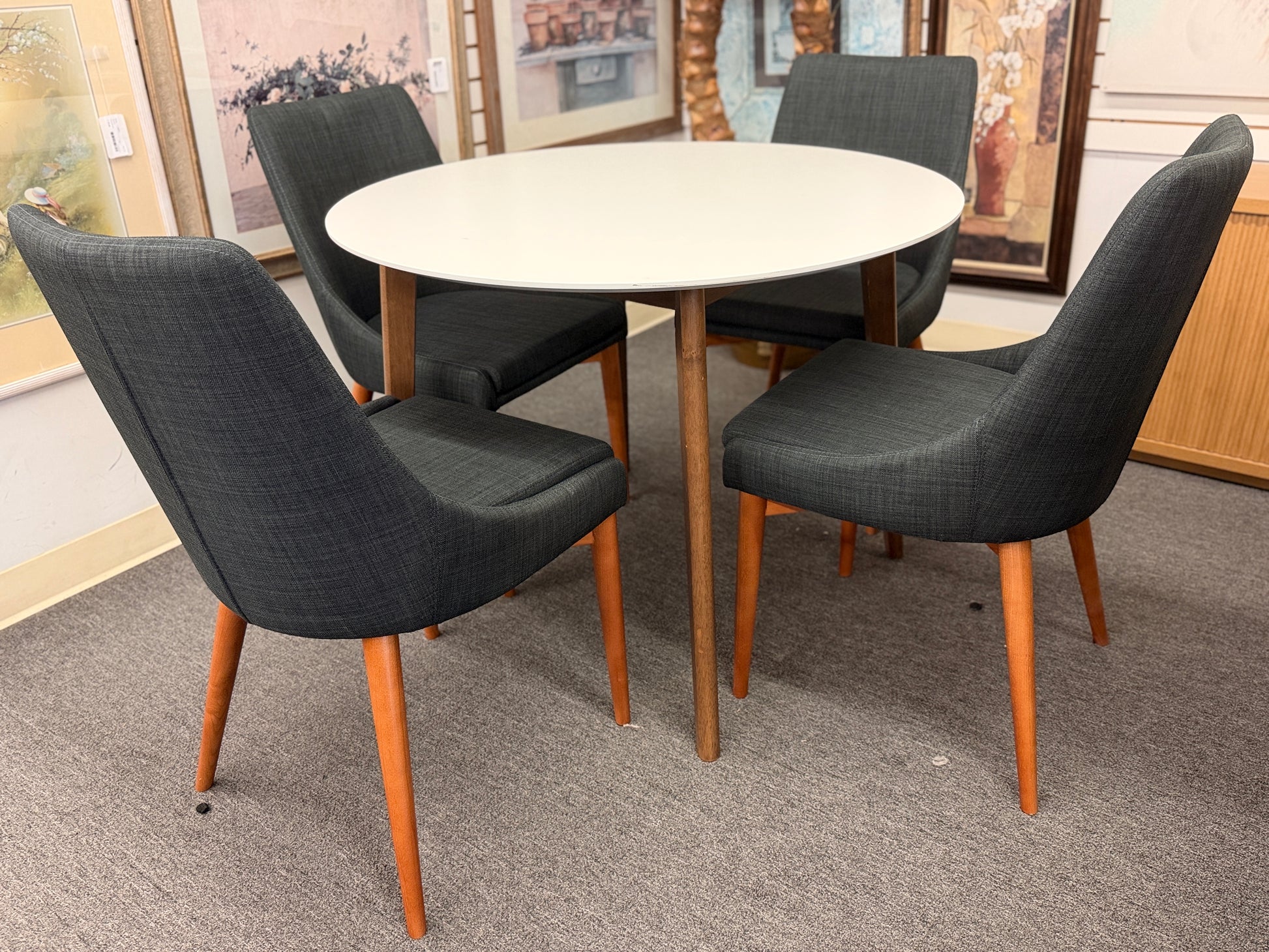 A mid-century modern dining set featuring a round white tabletop with tapered wooden legs and four dark gray upholstered chairs.
