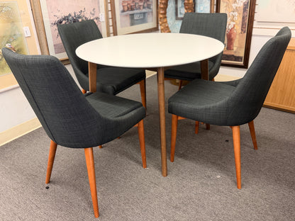 A mid-century modern dining set featuring a round white tabletop with tapered wooden legs and four dark gray upholstered chairs.