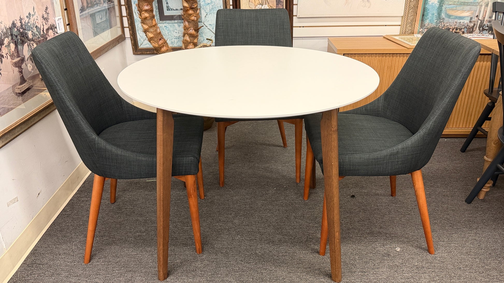 A mid-century modern dining set featuring a round white tabletop with tapered wooden legs and four dark gray upholstered chairs.