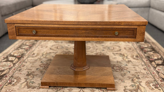 A mid-century inspired square wooden coffee table featuring a parquet veneer top, single drawer with antique brass pulls, and a central pedestal on a flat plinth base in good condition.