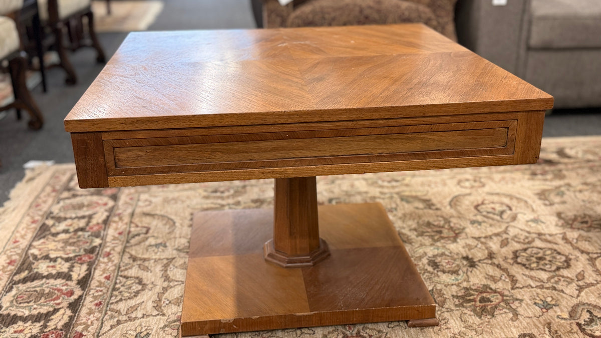 A mid-century inspired square wooden coffee table featuring a parquet veneer top, single drawer with antique brass pulls, and a central pedestal on a flat plinth base in good condition.