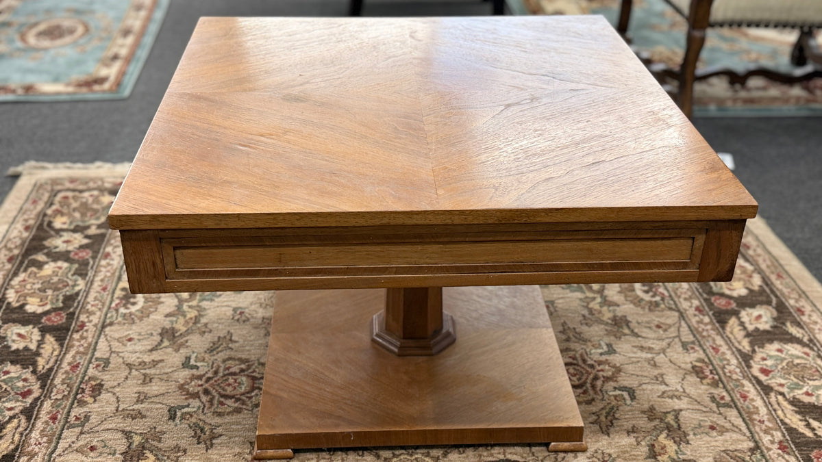 A mid-century inspired square wooden coffee table featuring a parquet veneer top, single drawer with antique brass pulls, and a central pedestal on a flat plinth base in good condition.