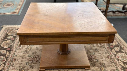 A mid-century inspired square wooden coffee table featuring a parquet veneer top, single drawer with antique brass pulls, and a central pedestal on a flat plinth base in good condition.