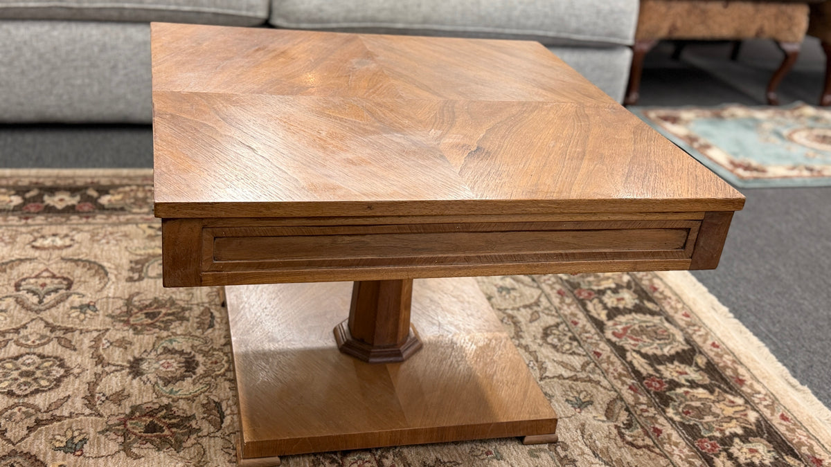 A mid-century inspired square wooden coffee table featuring a parquet veneer top, single drawer with antique brass pulls, and a central pedestal on a flat plinth base in good condition.