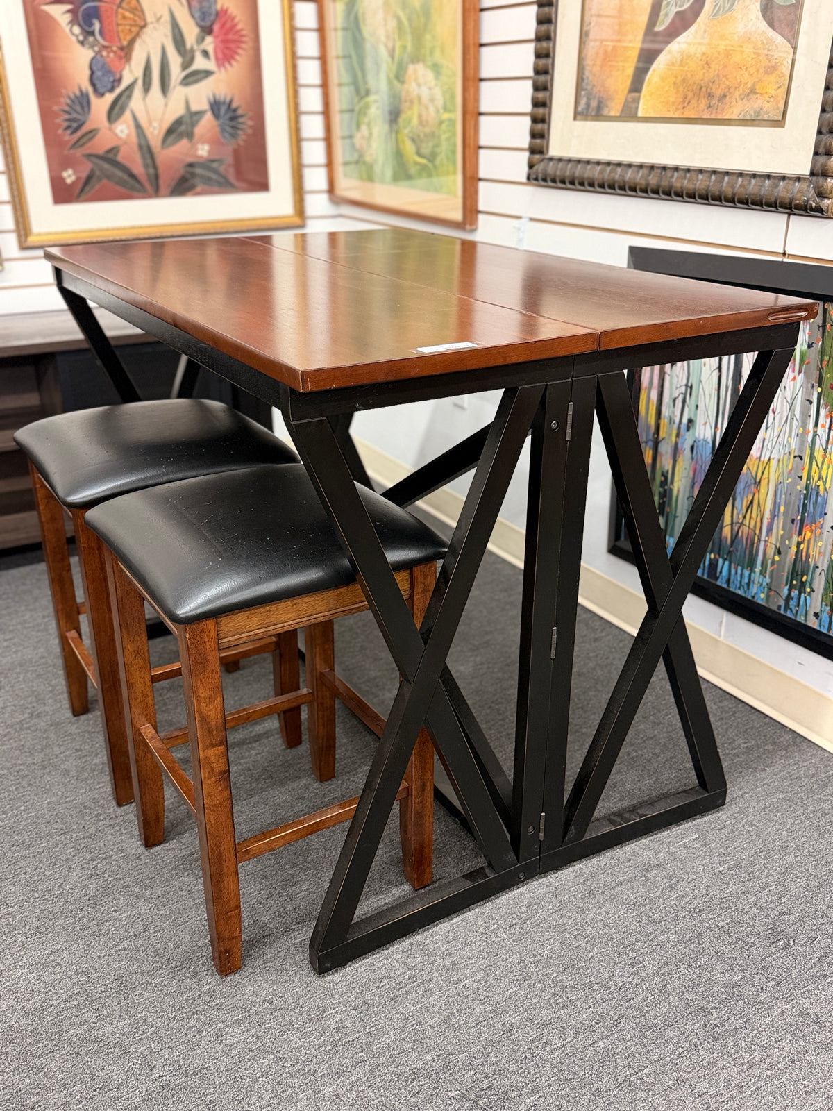 A counter-height dining set featuring a warm wood drop-leaf top, black metal X-frame base, and two matching backless barstools with black faux-leather cushioned seats in good condition.