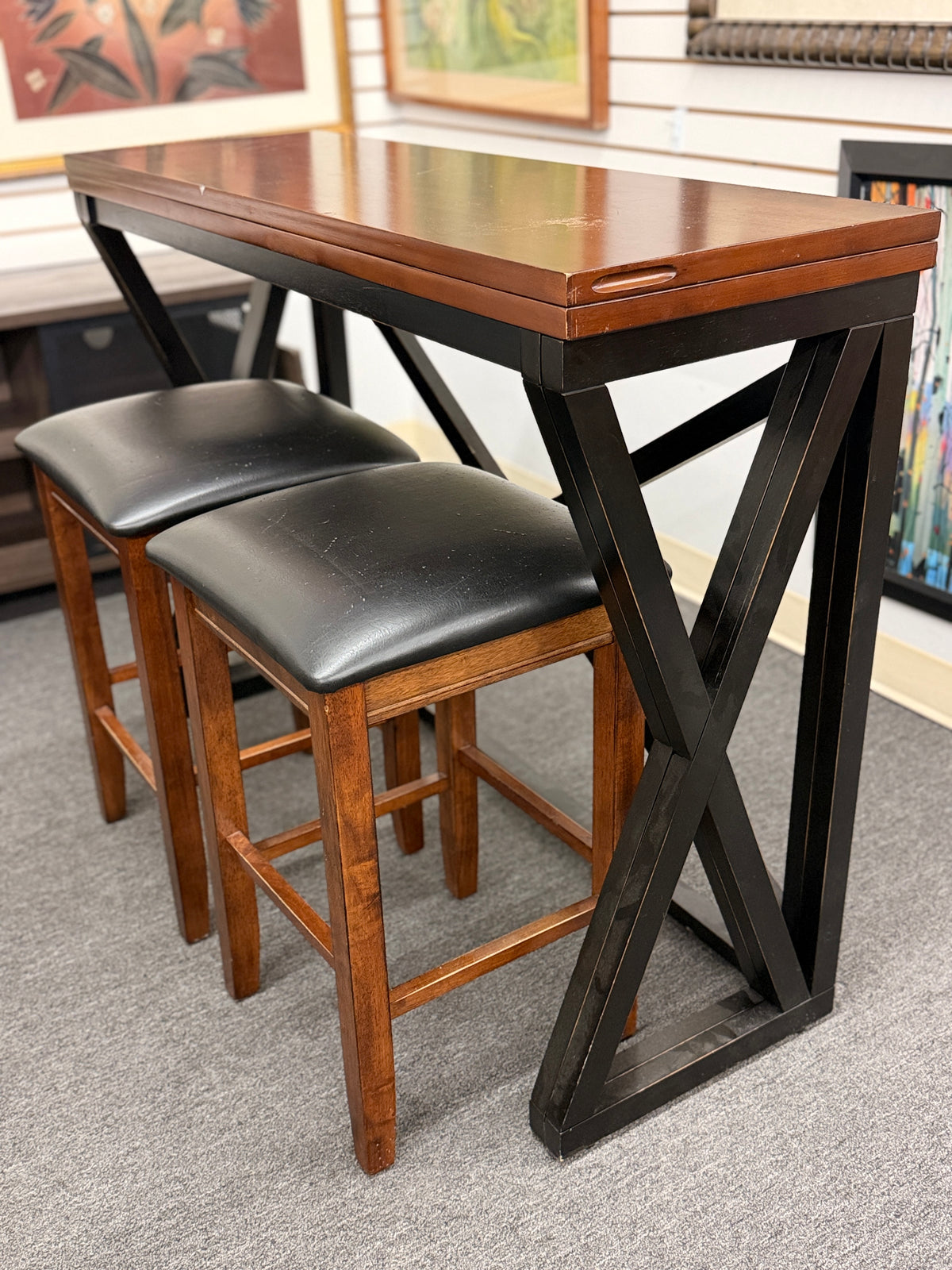 A counter-height dining set featuring a warm wood drop-leaf top, black metal X-frame base, and two matching backless barstools with black faux-leather cushioned seats in good condition.