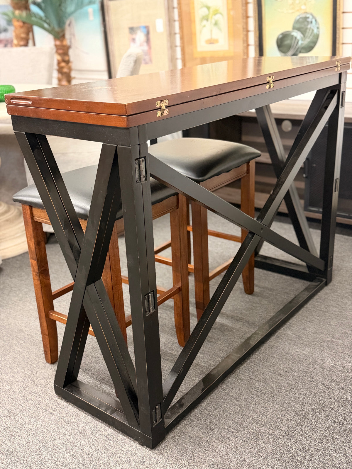 A counter-height dining set featuring a warm wood drop-leaf top, black metal X-frame base, and two matching backless barstools with black faux-leather cushioned seats in good condition.