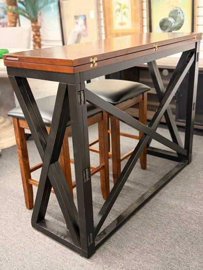 A counter-height dining set featuring a warm wood drop-leaf top, black metal X-frame base, and two matching backless barstools with black faux-leather cushioned seats in good condition.