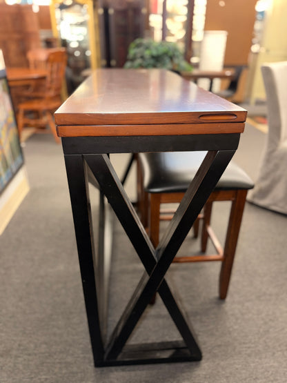 A counter-height dining set featuring a warm wood drop-leaf top, black metal X-frame base, and two matching backless barstools with black faux-leather cushioned seats in good condition.