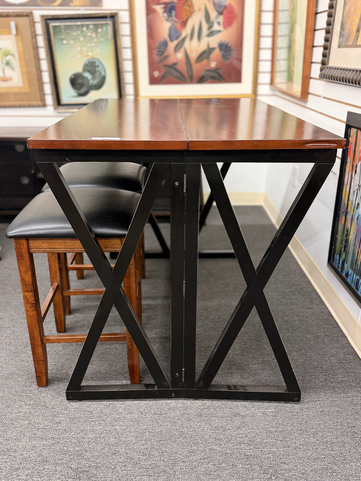 A counter-height dining set featuring a warm wood drop-leaf top, black metal X-frame base, and two matching backless barstools with black faux-leather cushioned seats in good condition.