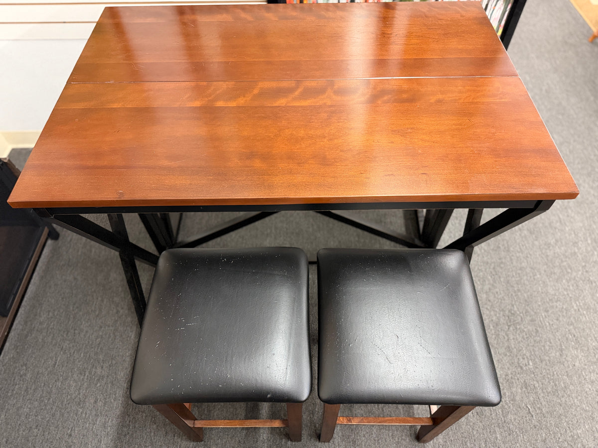 A counter-height dining set featuring a warm wood drop-leaf top, black metal X-frame base, and two matching backless barstools with black faux-leather cushioned seats in good condition.