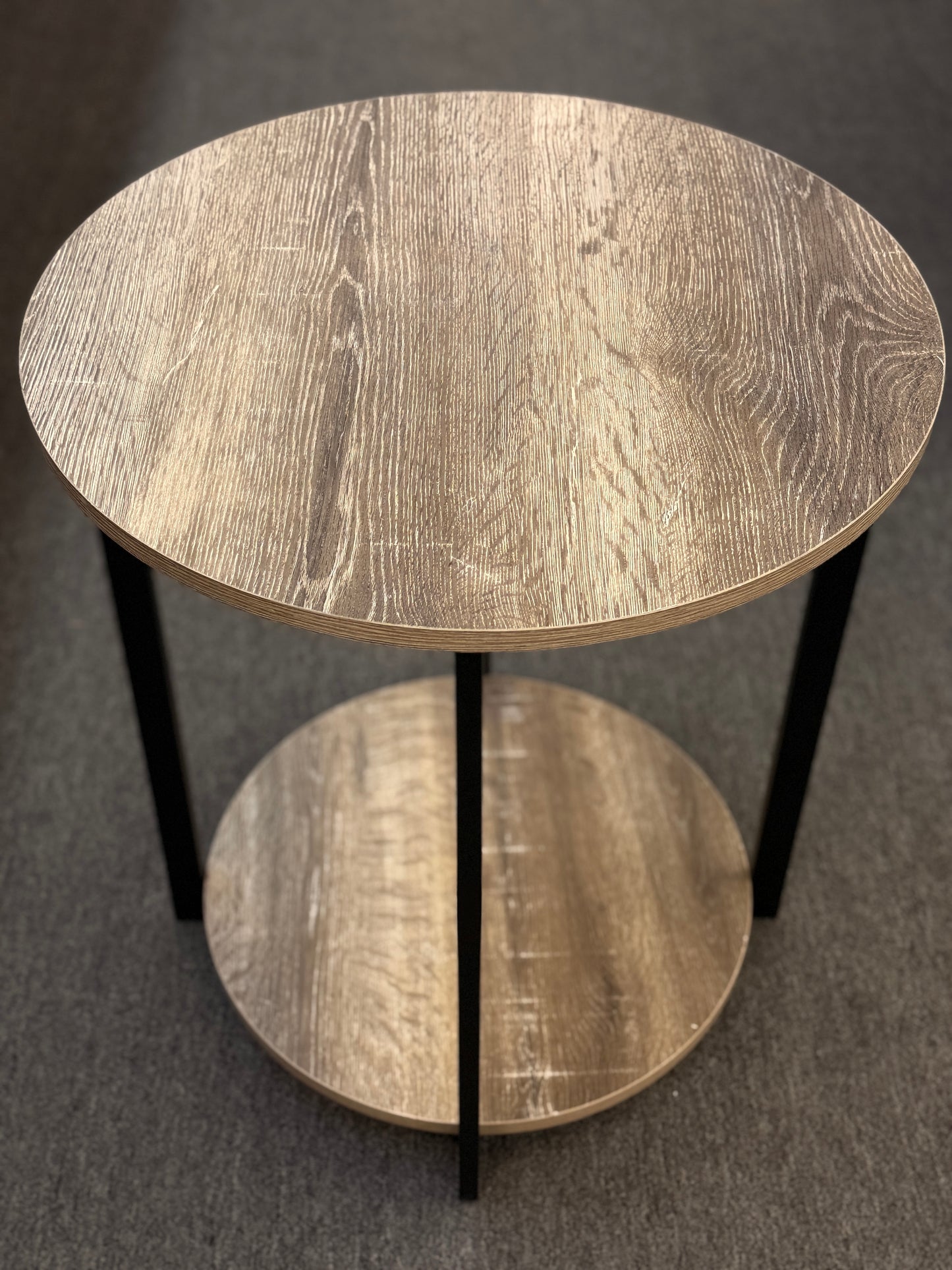 A round two-tiered end table featuring a distressed faux wood-grain finish on both circular shelves with a minimalist black iron frame.
