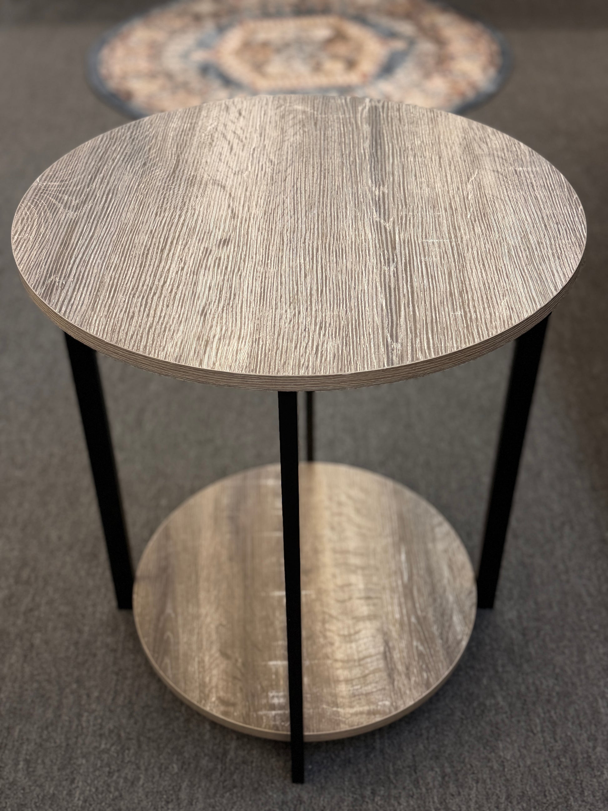 A round two-tiered end table featuring a distressed faux wood-grain finish on both circular shelves with a minimalist black iron frame.