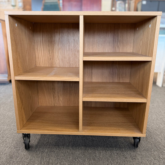 A modern mobile office storage cart with a light oak finish, featuring asymmetrical divided cubby compartments and four black rolling casters.