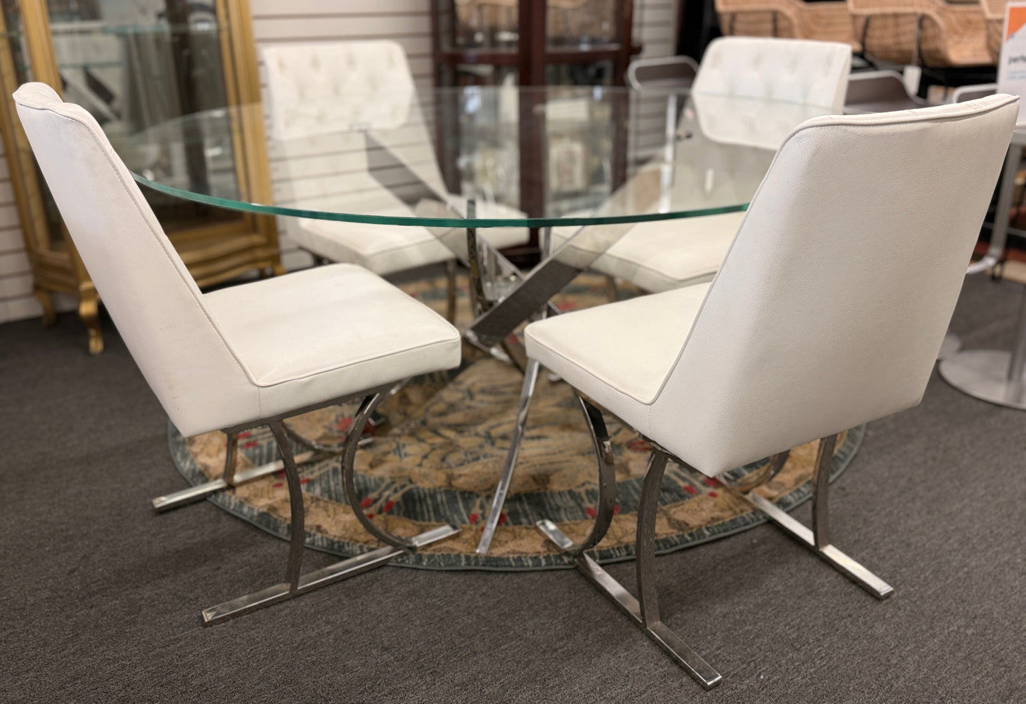 A modern dining set featuring a round glass-top table on an angled iron base with four white high-back tufted chairs.