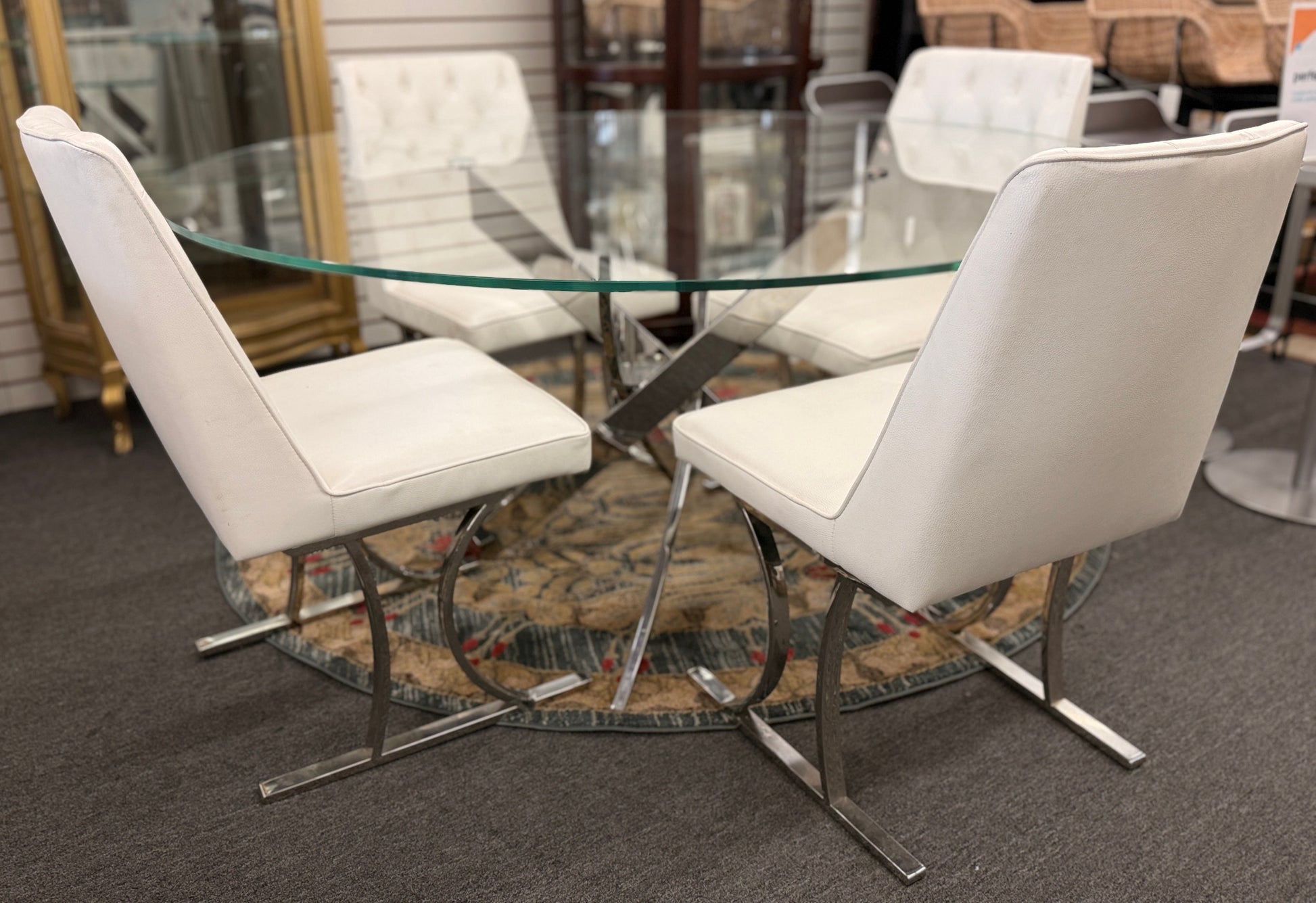 A modern dining set featuring a round glass-top table on an angled iron base with four white high-back tufted chairs.