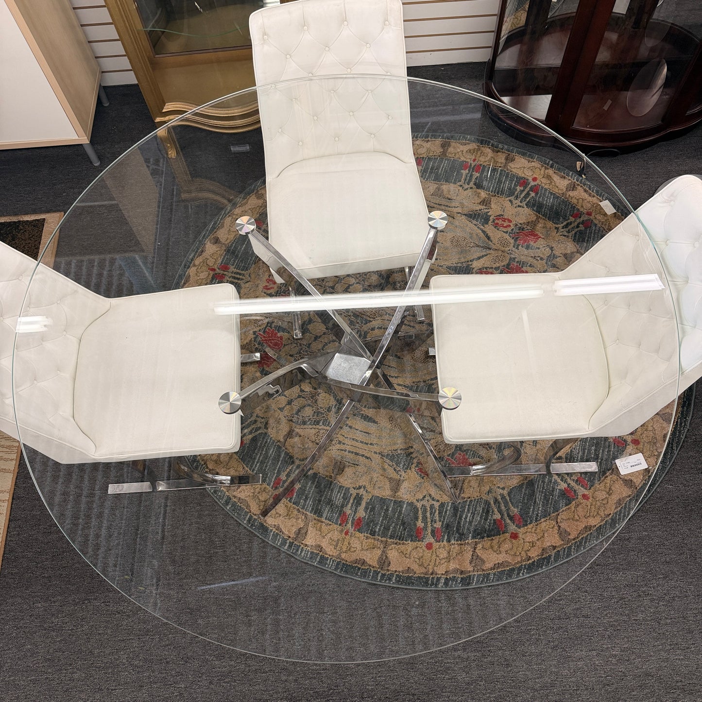A modern dining set featuring a round glass-top table on an angled iron base with four white high-back tufted chairs.