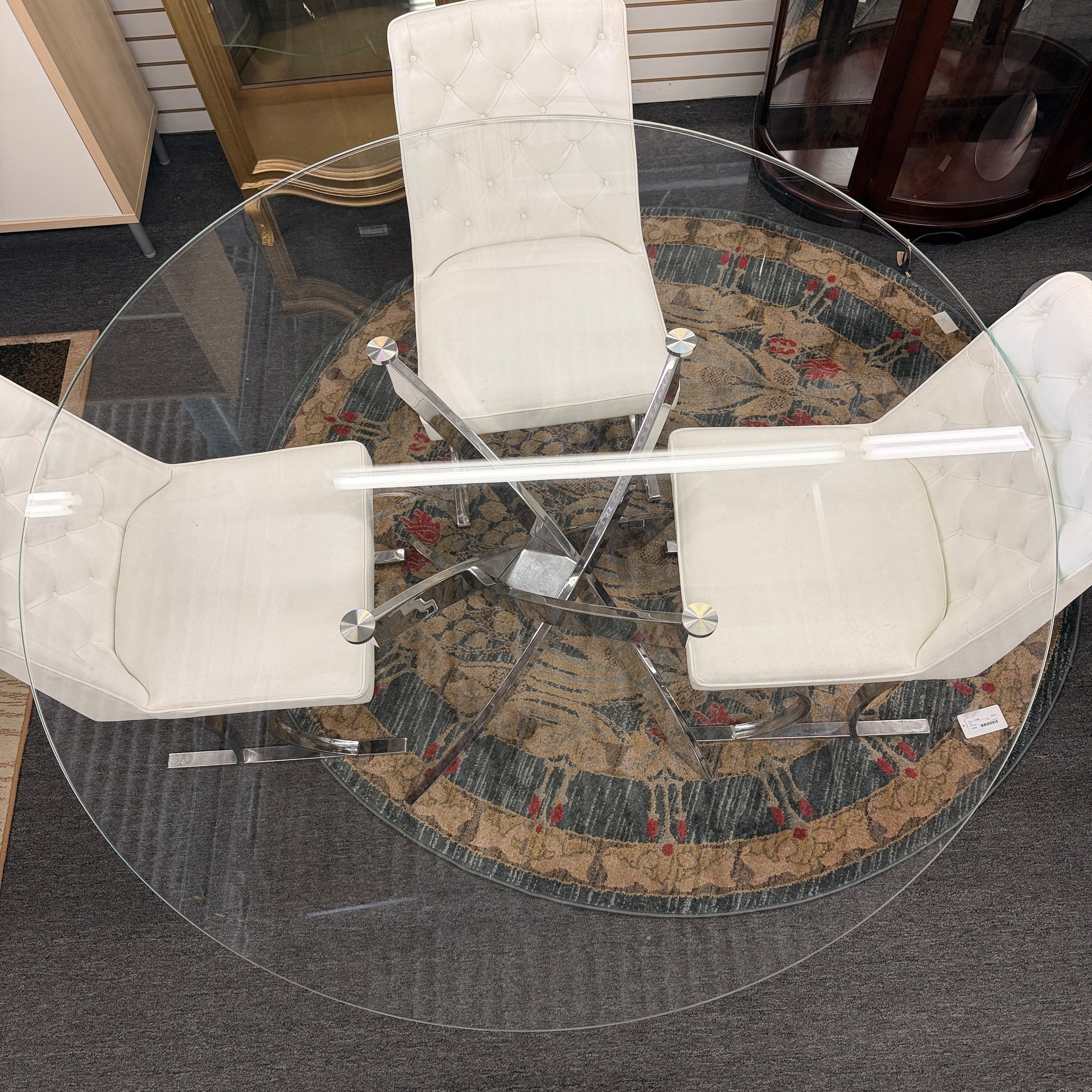 A modern dining set featuring a round glass-top table on an angled iron base with four white high-back tufted chairs.
