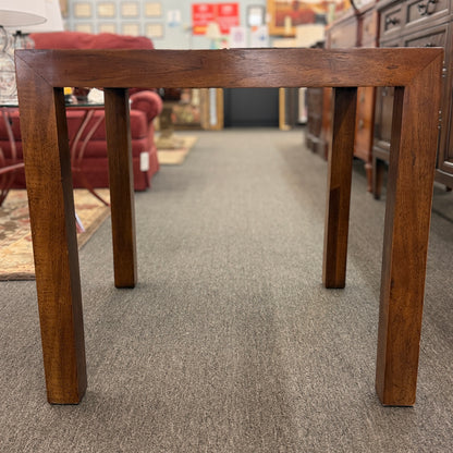 A modern square accent table with thick blocky legs flush with the corners and a highly reflective, glossy top surface showing a rich burl wood pattern.
