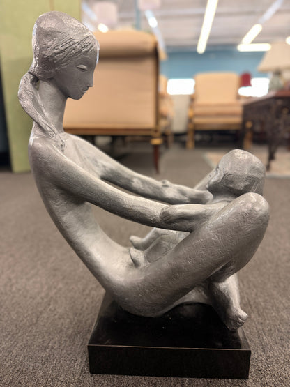A stylized modern sculpture of a seated mother holding a young child with smooth elongated lines and a textured metallic finish mounted on a solid block base.