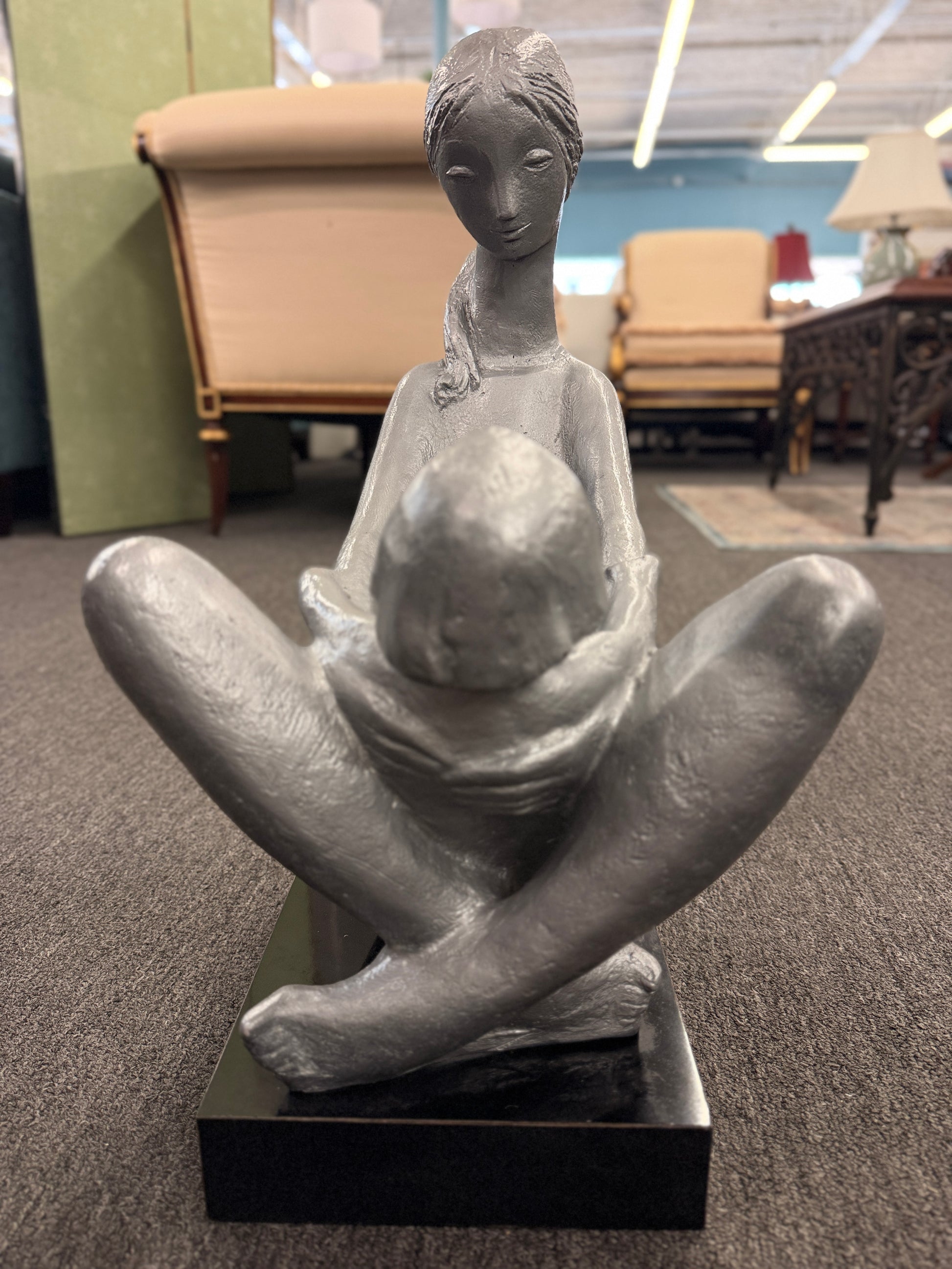 A stylized modern sculpture of a seated mother holding a young child with smooth elongated lines and a textured metallic finish mounted on a solid block base.