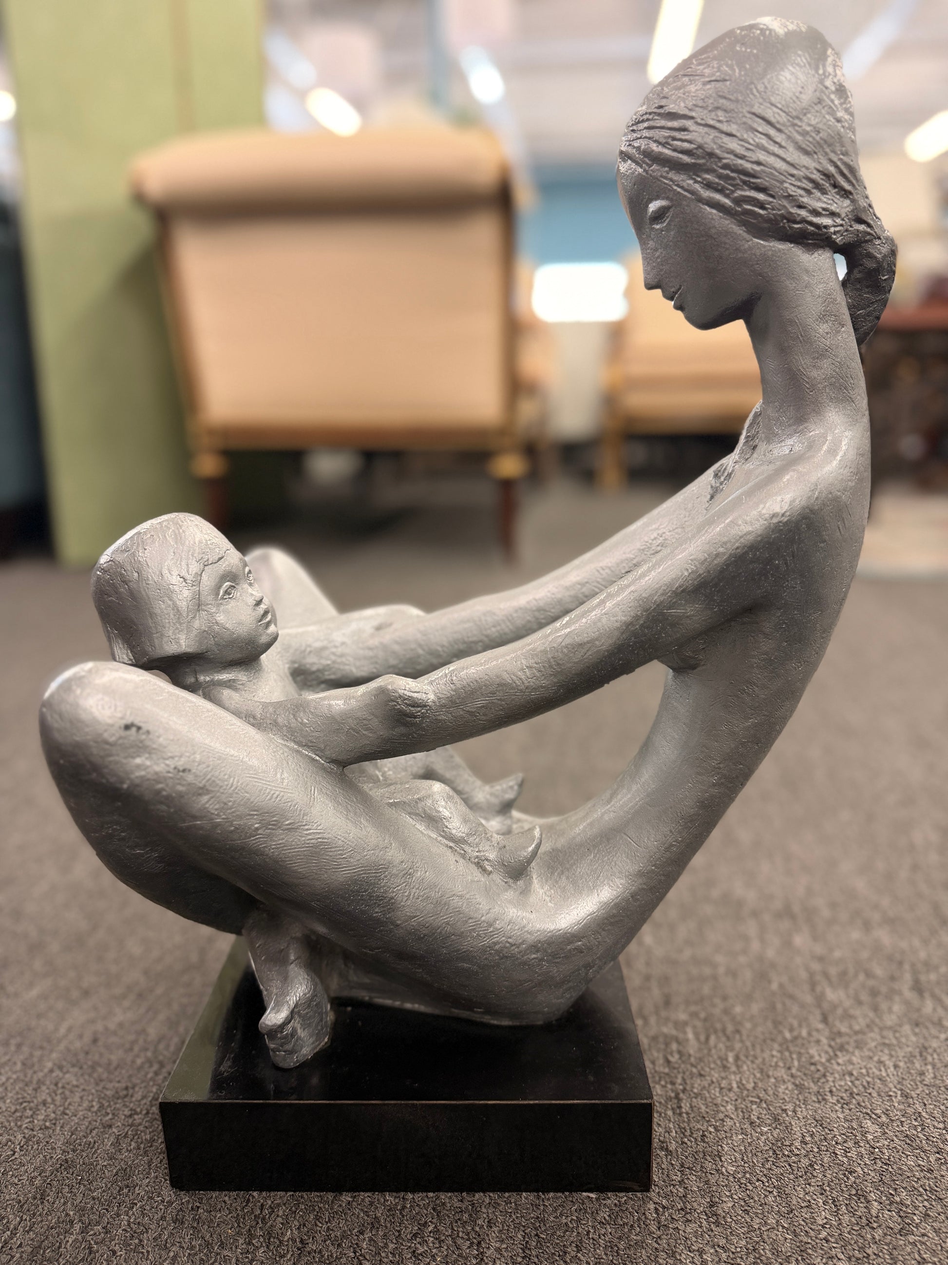 A stylized modern sculpture of a seated mother holding a young child with smooth elongated lines and a textured metallic finish mounted on a solid block base.