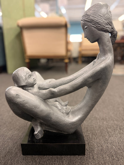A stylized modern sculpture of a seated mother holding a young child with smooth elongated lines and a textured metallic finish mounted on a solid block base.