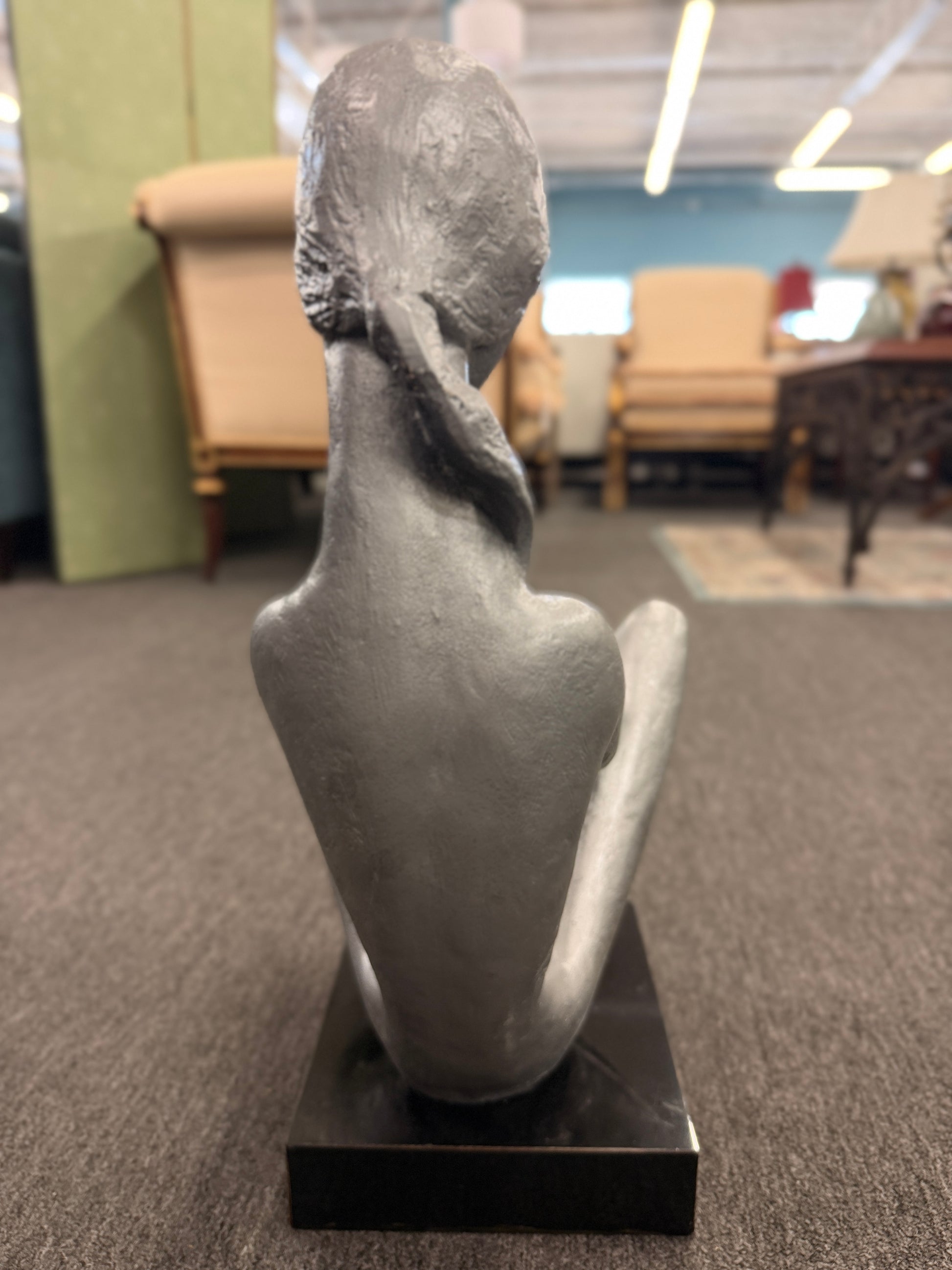 A stylized modern sculpture of a seated mother holding a young child with smooth elongated lines and a textured metallic finish mounted on a solid block base.