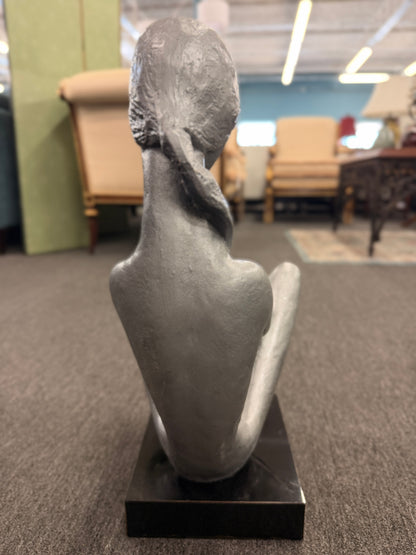 A stylized modern sculpture of a seated mother holding a young child with smooth elongated lines and a textured metallic finish mounted on a solid block base.