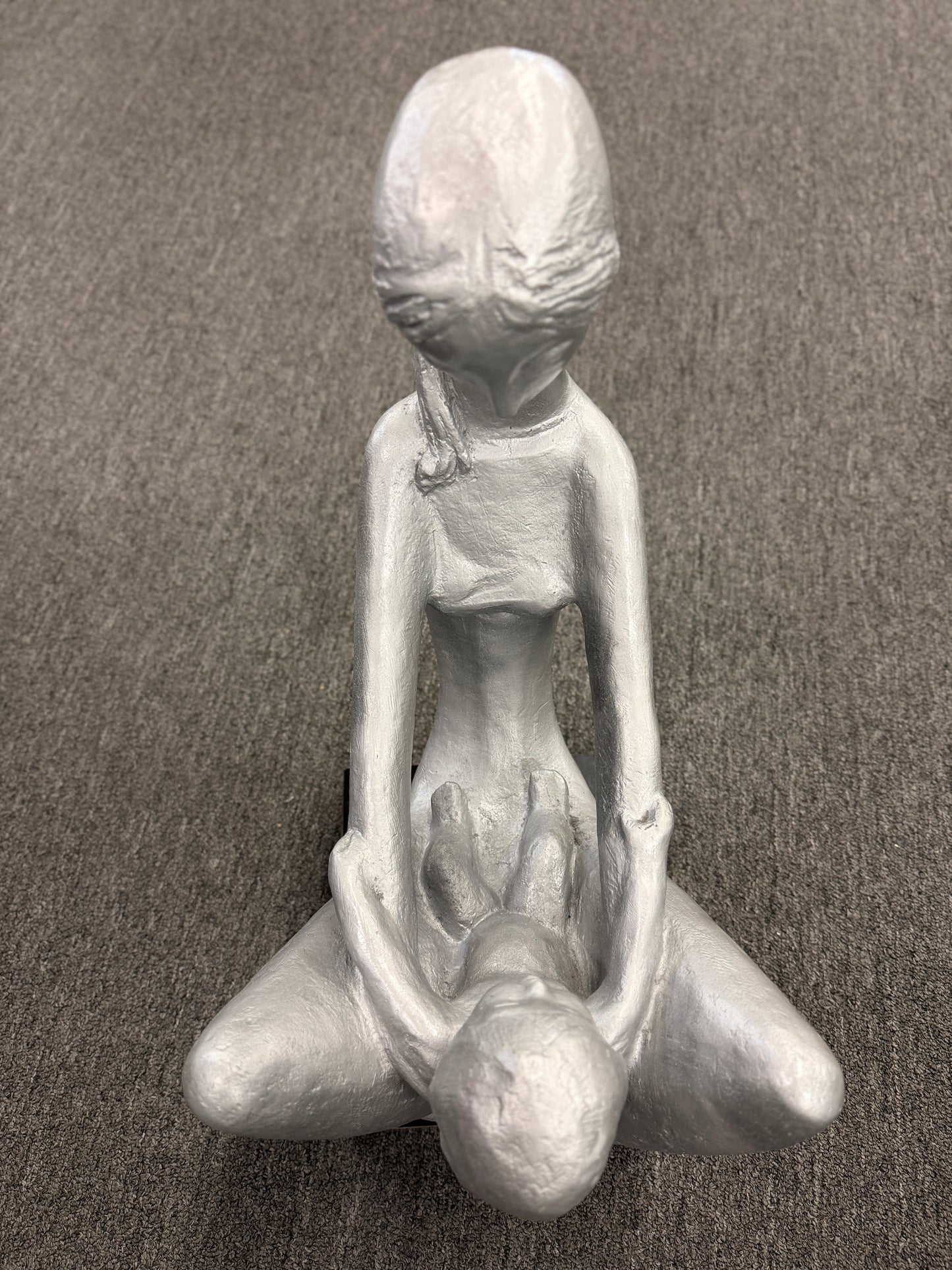 A stylized modern sculpture of a seated mother holding a young child with smooth elongated lines and a textured metallic finish mounted on a solid block base.