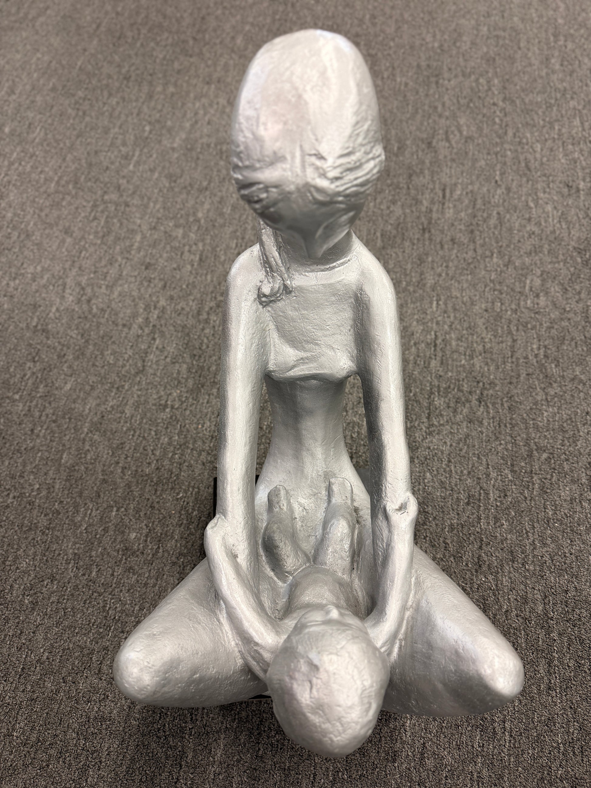 A stylized modern sculpture of a seated mother holding a young child with smooth elongated lines and a textured metallic finish mounted on a solid block base.