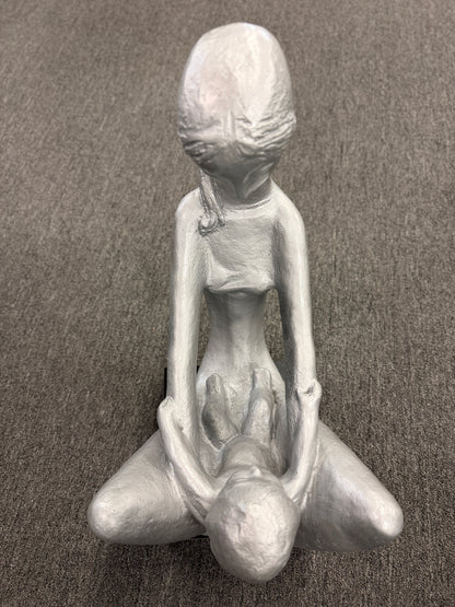 A stylized modern sculpture of a seated mother holding a young child with smooth elongated lines and a textured metallic finish mounted on a solid block base.