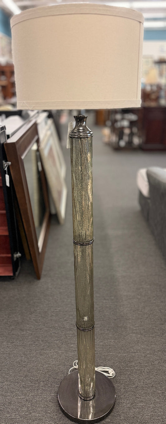A tall floor lamp featuring a slender textured metallic column base and a neutral drum shade in excellent condition.