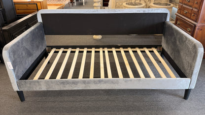 A modern daybed frame upholstered in a gray textured fabric, featuring a mid-height backrest, enclosed side panels, and a wooden slatted mattress support base.