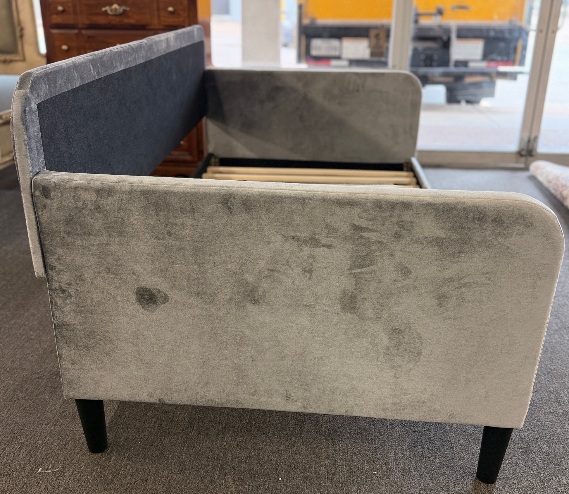 A modern daybed frame upholstered in a gray textured fabric, featuring a mid-height backrest, enclosed side panels, and a wooden slatted mattress support base.