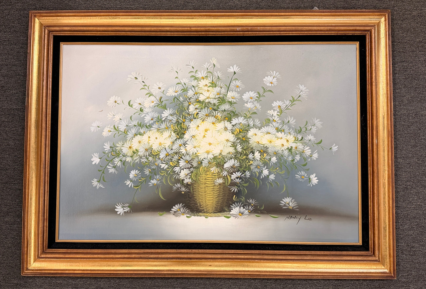 A traditional still-life canvas painting by Nancy Lee showing a dense bouquet of white daisies in a wicker basket, set in a dark velvet liner and an ornate gilded gold frame.
