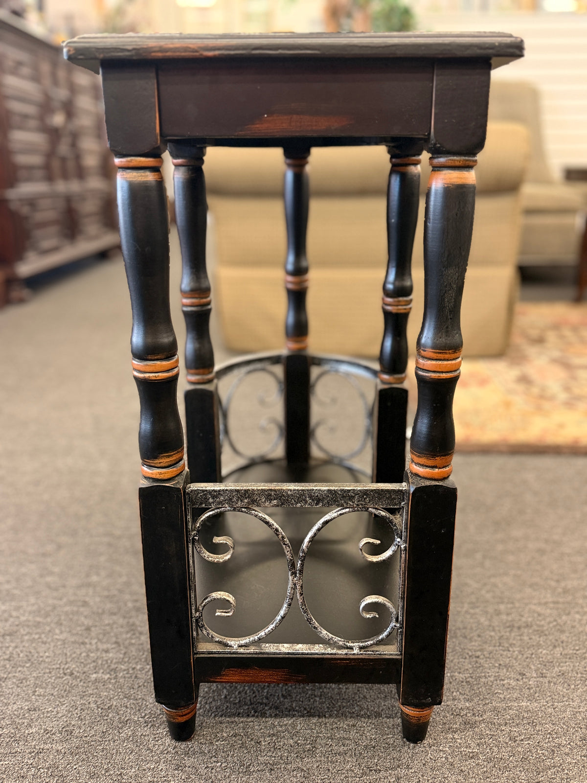 A narrow wooden accent table and magazine rack in a distressed black finish featuring turned spindle legs and scrolled metal side rails in good condition.