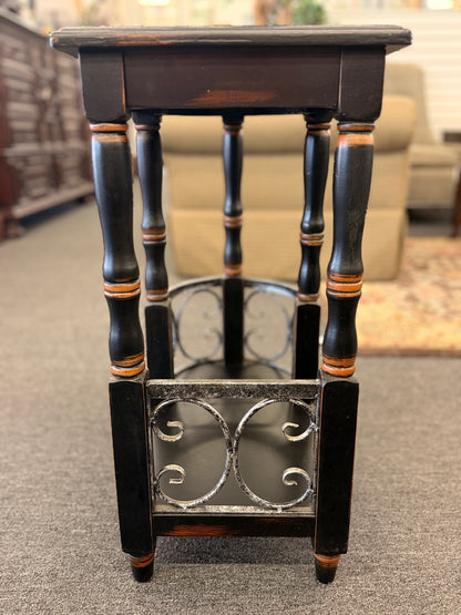 A narrow wooden accent table and magazine rack in a distressed black finish featuring turned spindle legs and scrolled metal side rails in good condition.