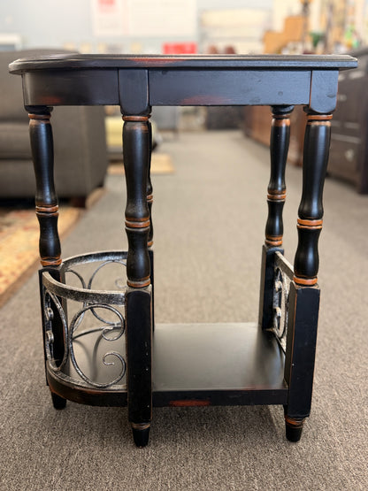 A narrow wooden accent table and magazine rack in a distressed black finish featuring turned spindle legs and scrolled metal side rails in good condition.