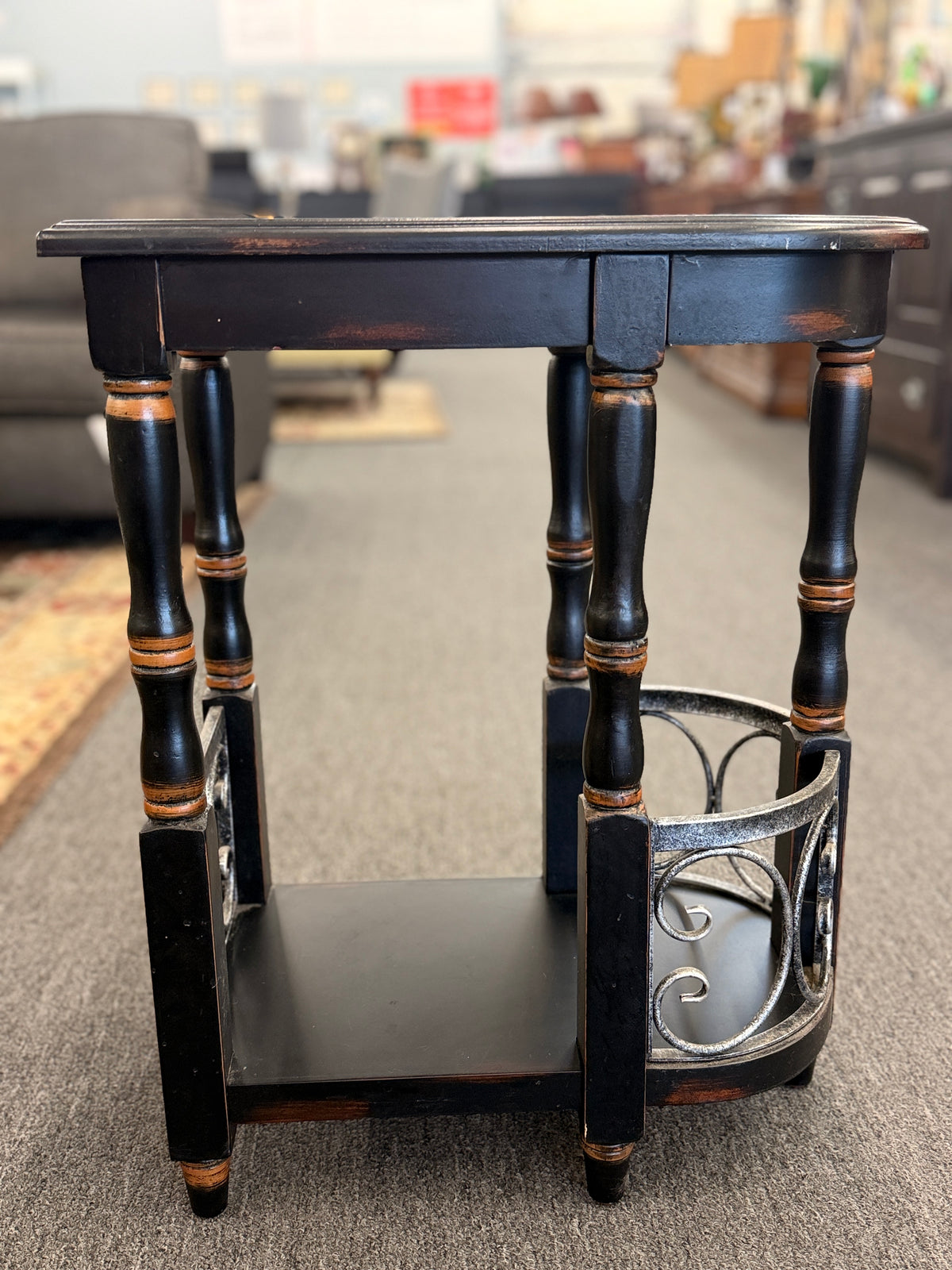 A narrow wooden accent table and magazine rack in a distressed black finish featuring turned spindle legs and scrolled metal side rails in good condition.