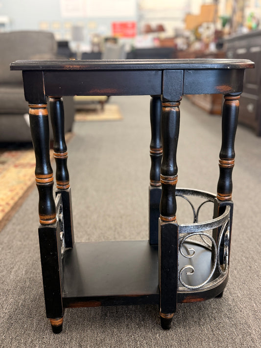 A narrow wooden accent table and magazine rack in a distressed black finish featuring turned spindle legs and scrolled metal side rails in good condition.