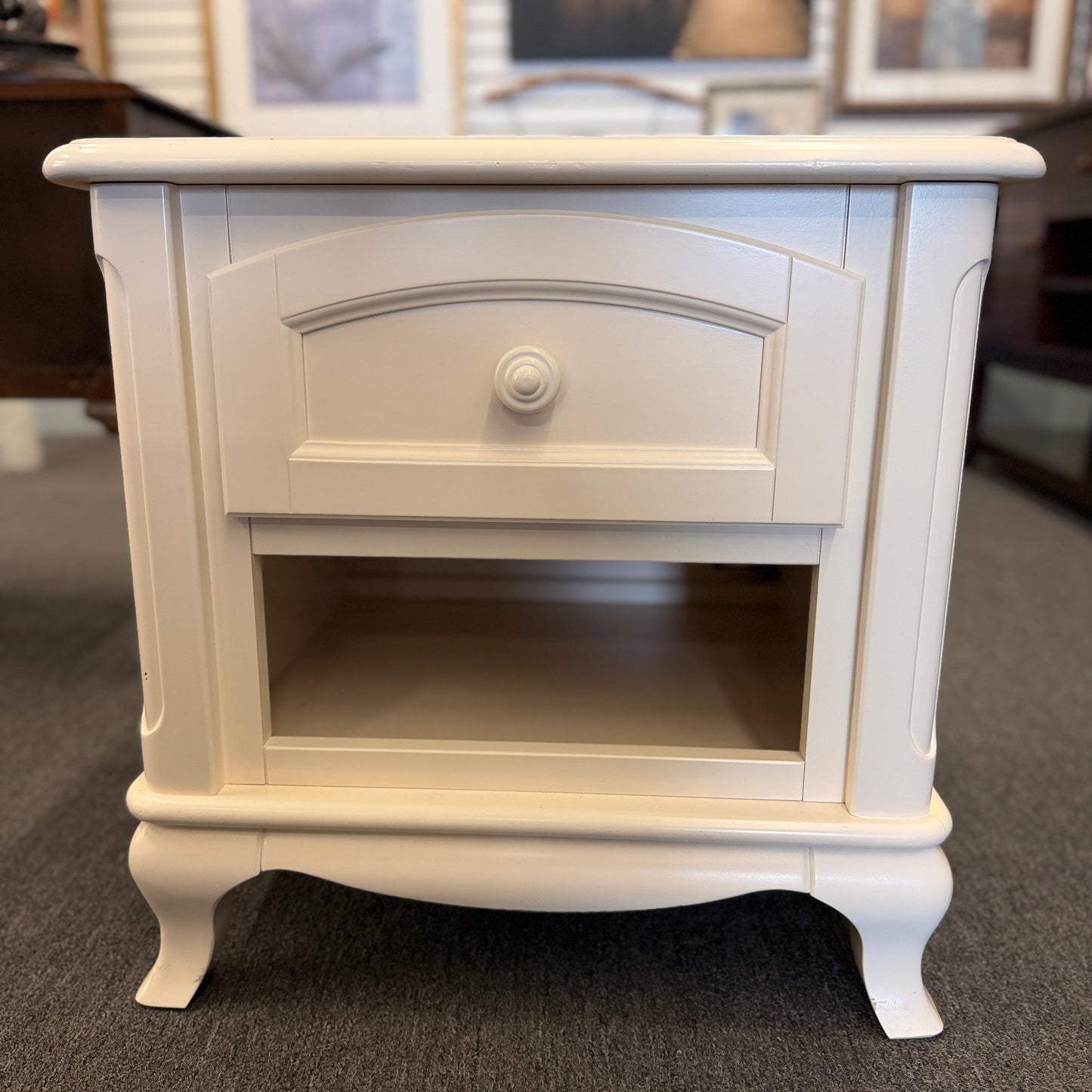 An off-white wooden nightstand featuring a single drawer with a turned knob, an open lower cubby, and curved cabriole legs.