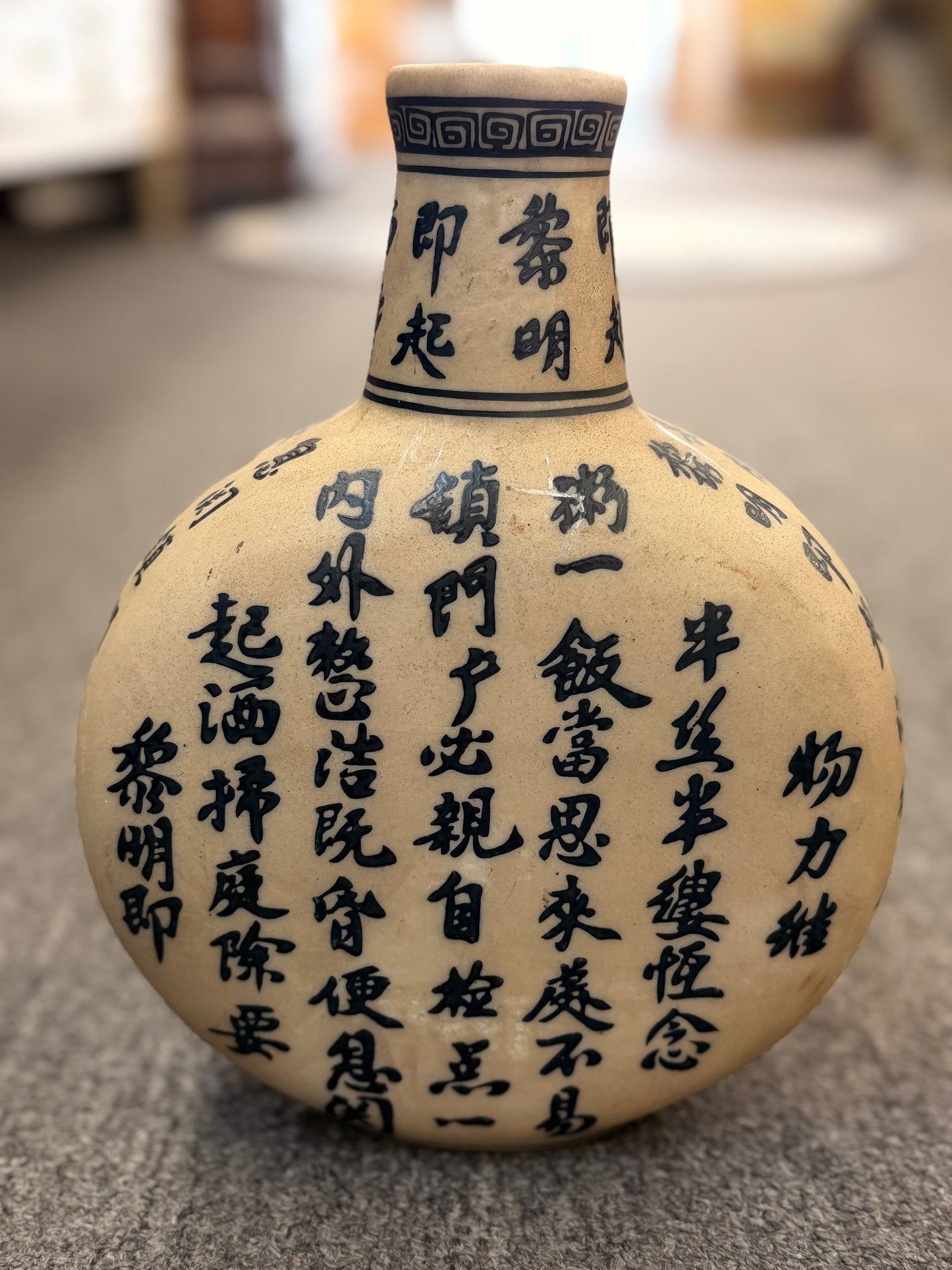 A round, flat-profile ceramic vase featuring black Asian calligraphy painted across a matte sand-colored surface.
